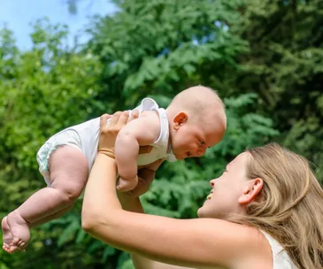 Mother joyfully lifting her smiling baby outdoors, surrounded by green trees.
