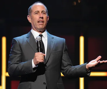 Comedian in a suit holding a microphone, gesturing on stage, with a blurred background.
