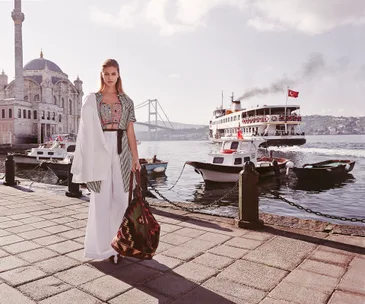 A woman in stylish attire stands by the Bosphorus in Istanbul, with the Ortaköy Mosque and a ferry in the background.