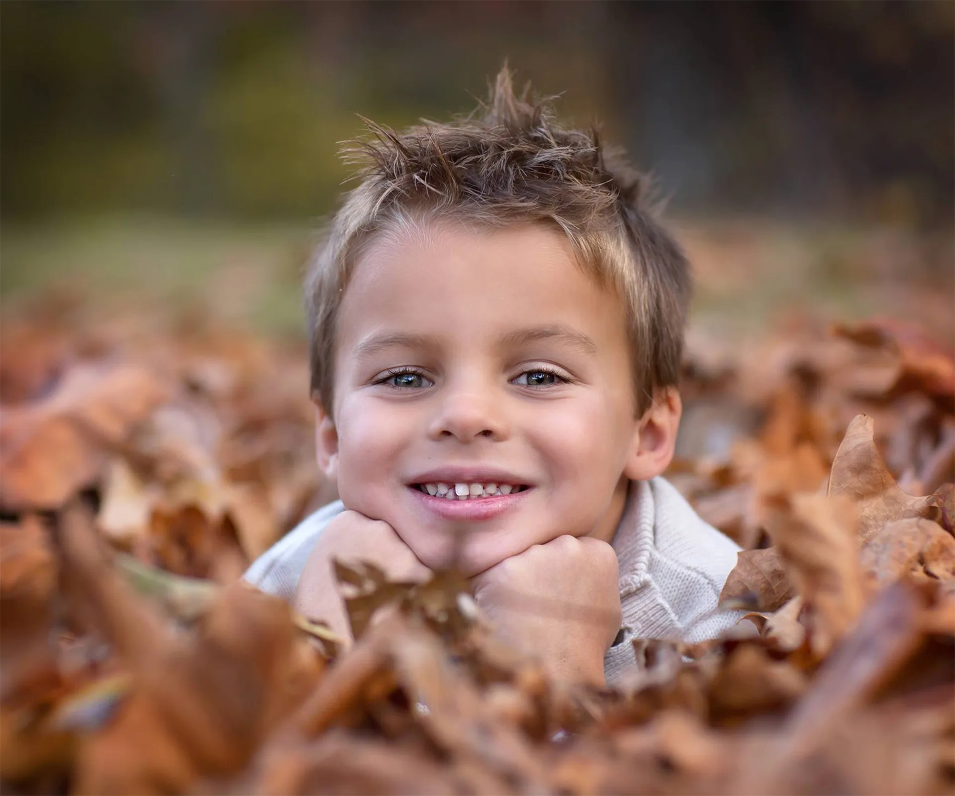 Young boy smiling, lying in a pile of autumn leaves, wearing a light sweater, with blurred background.