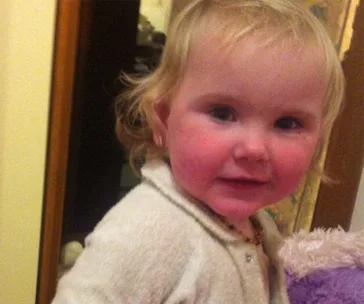 A young child with blonde hair looks towards the camera, wearing a light-colored outfit.