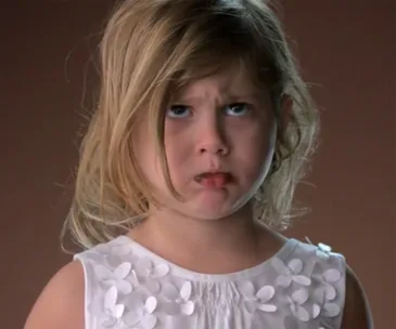 Young girl with blonde hair and a frowning expression, wearing a white dress with floral patterns against a plain background.