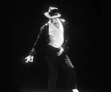 A performer in a fedora and sequined jacket strikes an iconic dance pose on stage against a dark background.