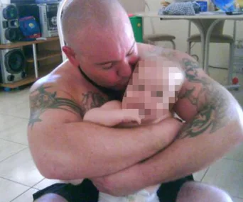 Shirtless man with tattoos hugging a baby, faces pixelated for privacy.