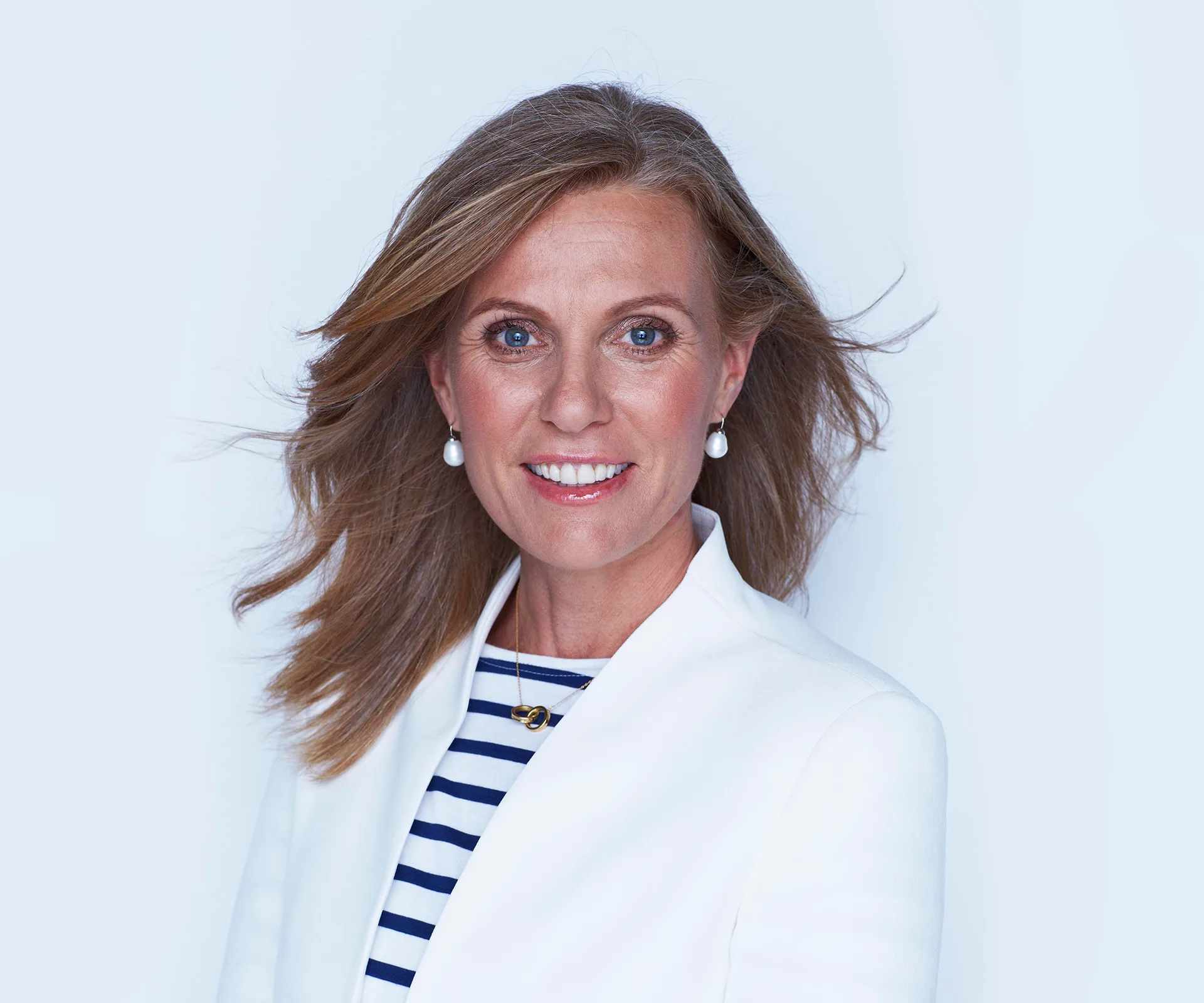 A person with long hair in a white jacket, wearing pearl earrings, smiles against a light background.