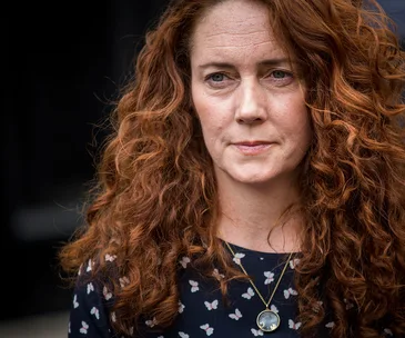 A person with long curly red hair, wearing a dark top with small pattern and a pendant necklace, looking ahead.