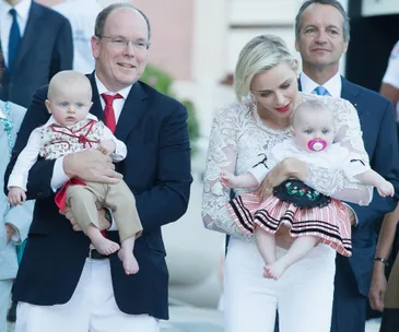 A smiling man and woman hold two babies, one in a suit and the other in a dress, with onlookers in the background.