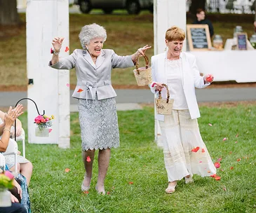 Two joyous grandmas in elegant outfits scatter flower petals at an outdoor wedding ceremony.