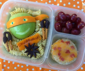 "Ninja Turtle-themed lunch with apple head, pasta, pretzel nunchucks, cheese pizza, and grapes on a polka dot cloth."
