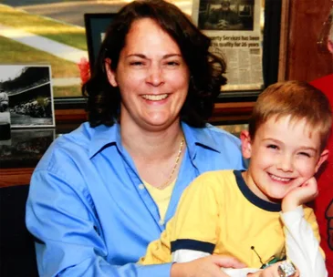 A woman in a blue shirt smiling, holding a young boy in a yellow shirt; framed photos in the background.