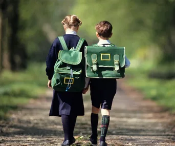 Two children in school uniforms walking on a path, wearing green backpacks.