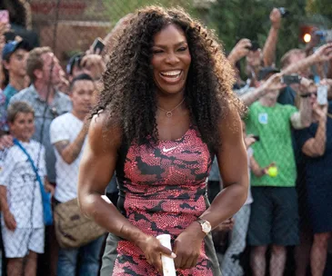 Tennis player smiling on a court, wearing a pink and black patterned outfit, surrounded by a cheering crowd.