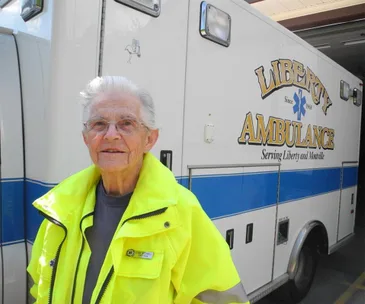 Elderly woman in a bright jacket stands in front of "Liberty Ambulance" with a smile.