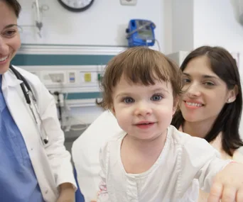 Toddler smiling in a hospital room with two women, including a doctor, indicating a supportive medical environment.