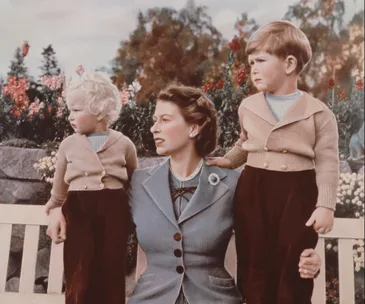 "A woman seated on a bench with two young children in matching outfits, surrounded by flowers in an outdoor setting."