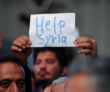 Man holds sign reading "Help Syria" at a gathering, symbolizing support for Syrian refugees.