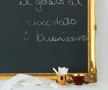 Vintage framed chalkboard with Italian writing, chalk pieces, and a red cup on a white table.