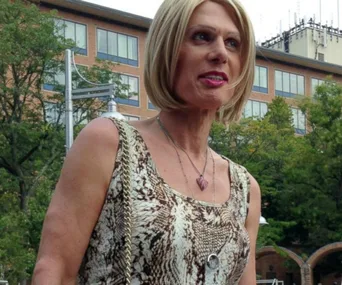 A woman in a sleeveless dress stands outdoors with trees and a building in the background, wearing a necklace.