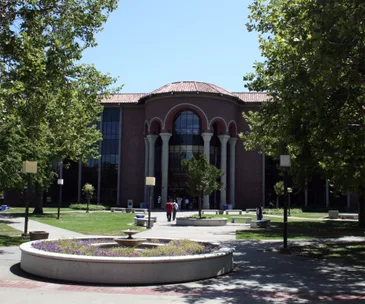 A large school building with tall arches, surrounded by trees and a circular garden in front.