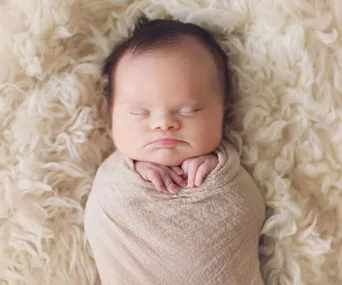 A peacefully sleeping baby wrapped in a beige blanket, lying on a soft, fluffy white surface.