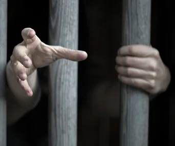 Hands reaching out through metal bars in a dimly lit setting, suggesting confinement.