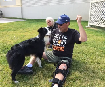 Man with leg brace sits on grass, petting a black and white dog; woman watches in background.