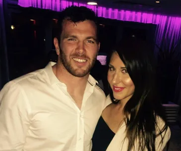A smiling man and woman posing together in a dimly lit room with purple lighting in the background.