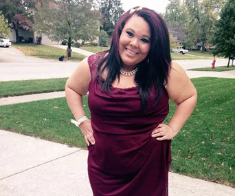 A smiling woman stands outdoors, wearing a burgundy dress, with hands on hips, on a suburban sidewalk.