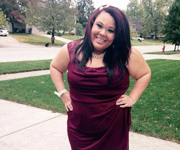 A smiling woman stands outdoors, wearing a burgundy dress, with hands on hips, on a suburban sidewalk.