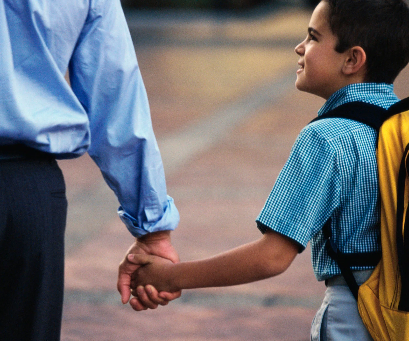 Adult and child holding hands, child wearing a blue checkered shirt and yellow backpack, walking outdoors.