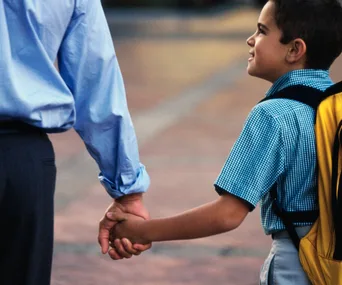 Adult and child holding hands, child wearing a blue checkered shirt and yellow backpack, walking outdoors.
