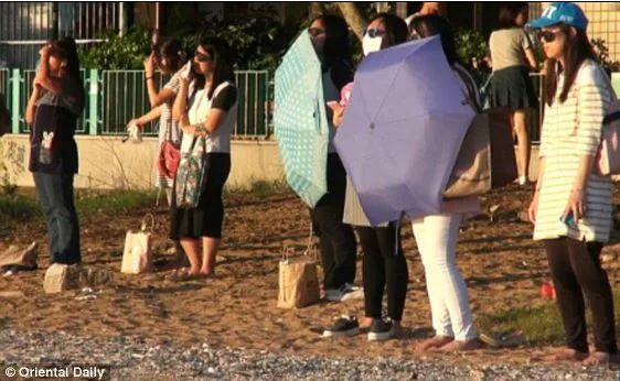 People standing on a beach, some holding umbrellas, observing the horizon during a sunset.