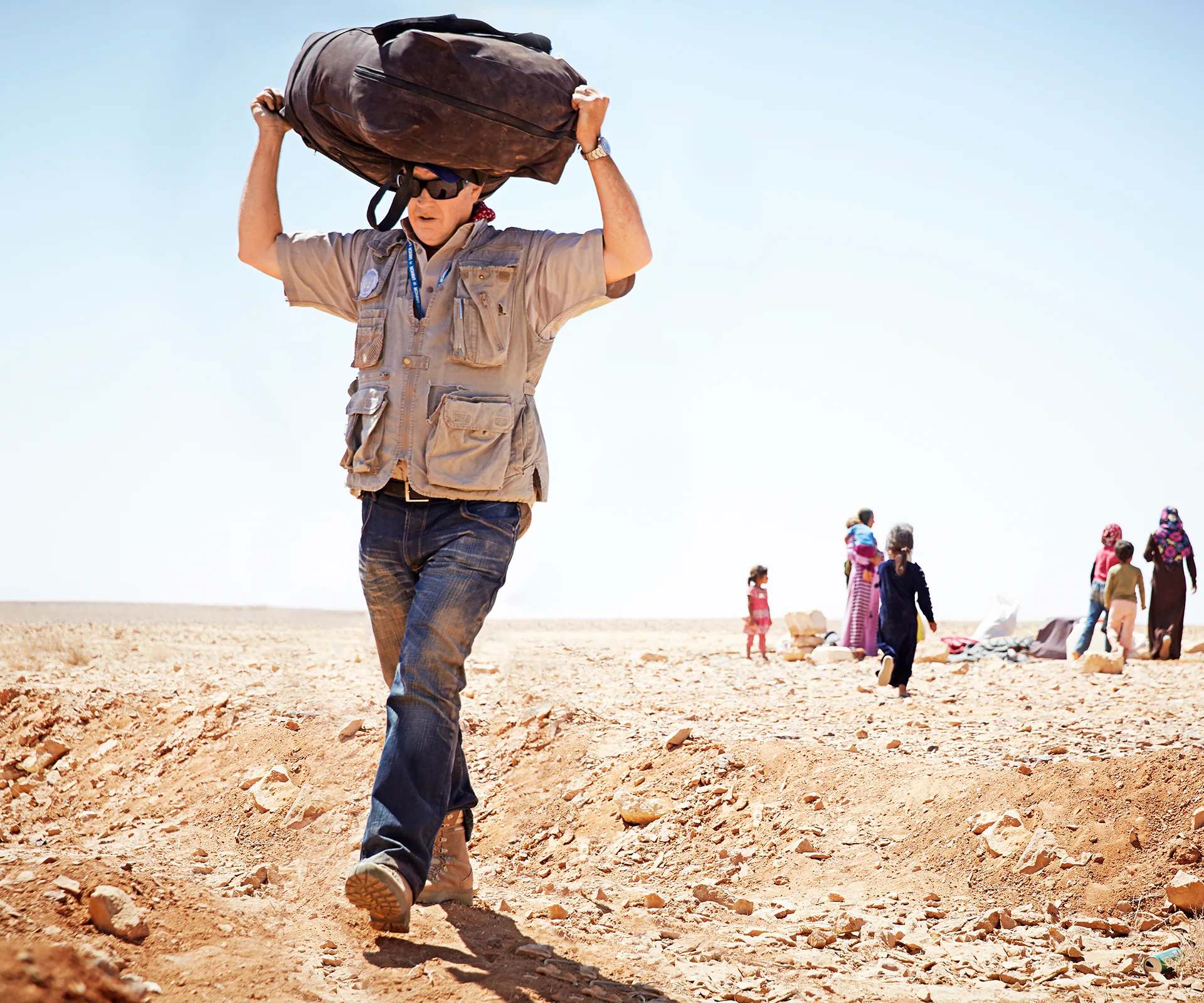 Man carrying bag on shoulders in desert; people in background, some children visible.