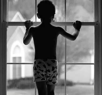 A child in patterned shorts stands indoors, silhouetted against a window, looking outside toward suburban houses.