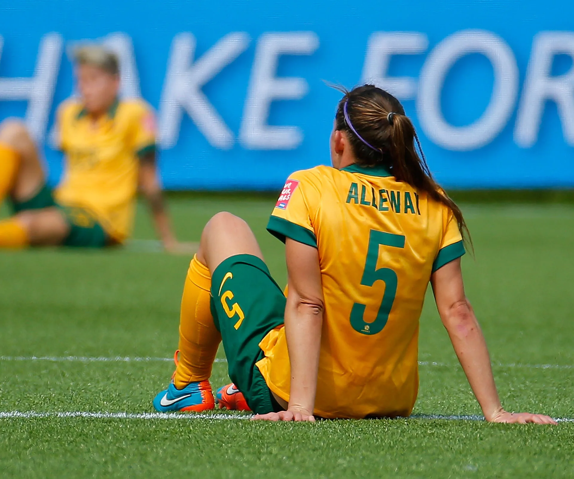 Australian soccer player sitting on the field, wearing a yellow jersey with "Alleway 5" on the back.