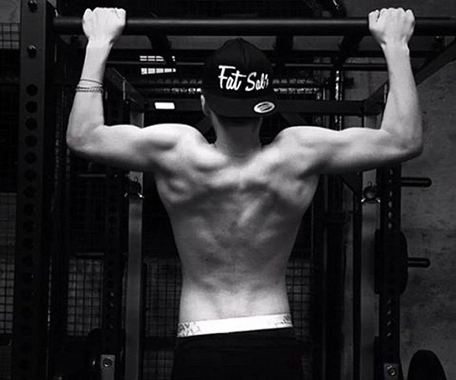 Back view of a man doing pull-ups, wearing a "Fat Sals" cap, showcasing toned muscles in a gym setting.