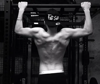 Back view of a man doing pull-ups, wearing a "Fat Sals" cap, showcasing toned muscles in a gym setting.
