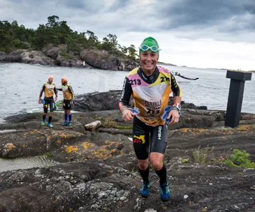Athlete in race gear runs on rocky shore during a triathlon event, with competitors and a kayak in the background.