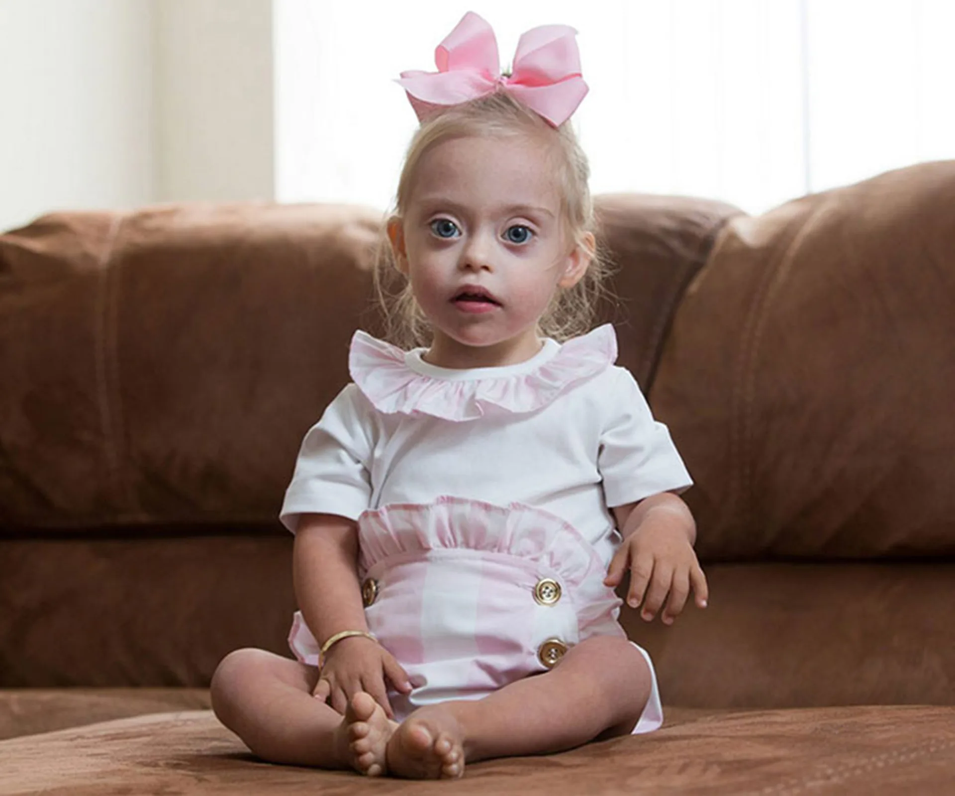 A toddler with Down syndrome, wearing a pink bow and white outfit, sits on a brown sofa.