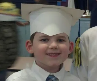 Young boy in a white graduation cap and shirt, smiling, with a tassel visible in the background.