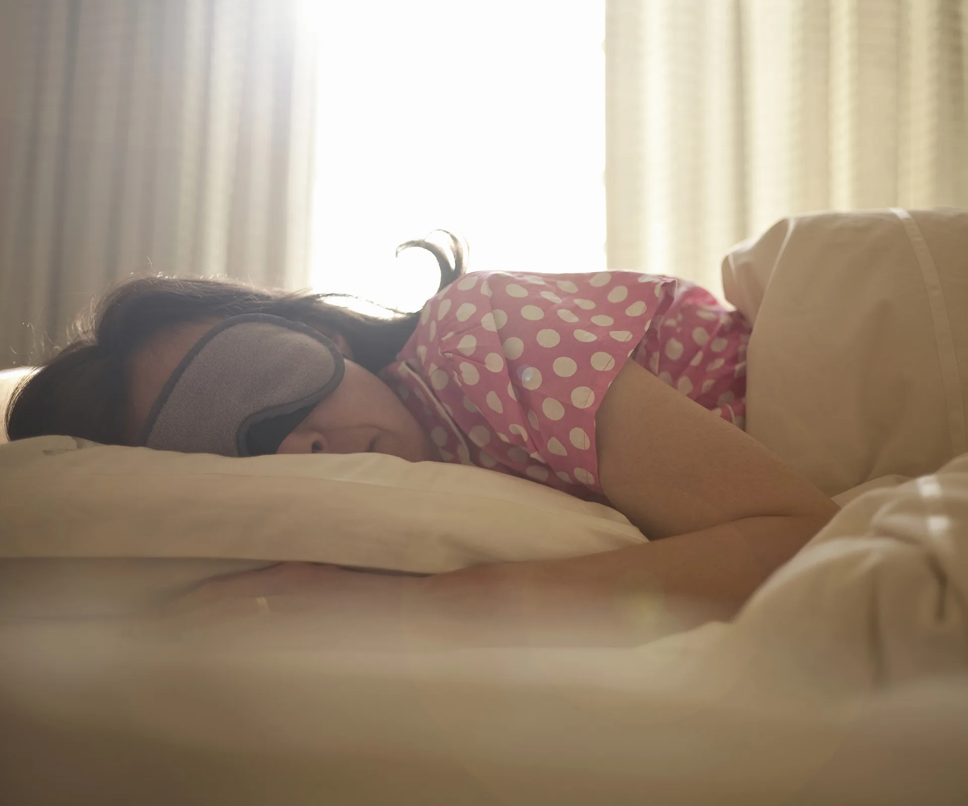 Woman in polka dot pajamas and sleep mask resting in bed, sunlight filtering through curtains.