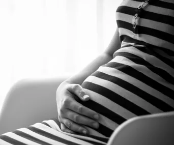 Pregnant woman in striped dress seated, hand gently resting on belly, black and white photo.