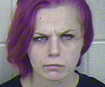 A person with purple hair and a serious expression, against a tiled wall.