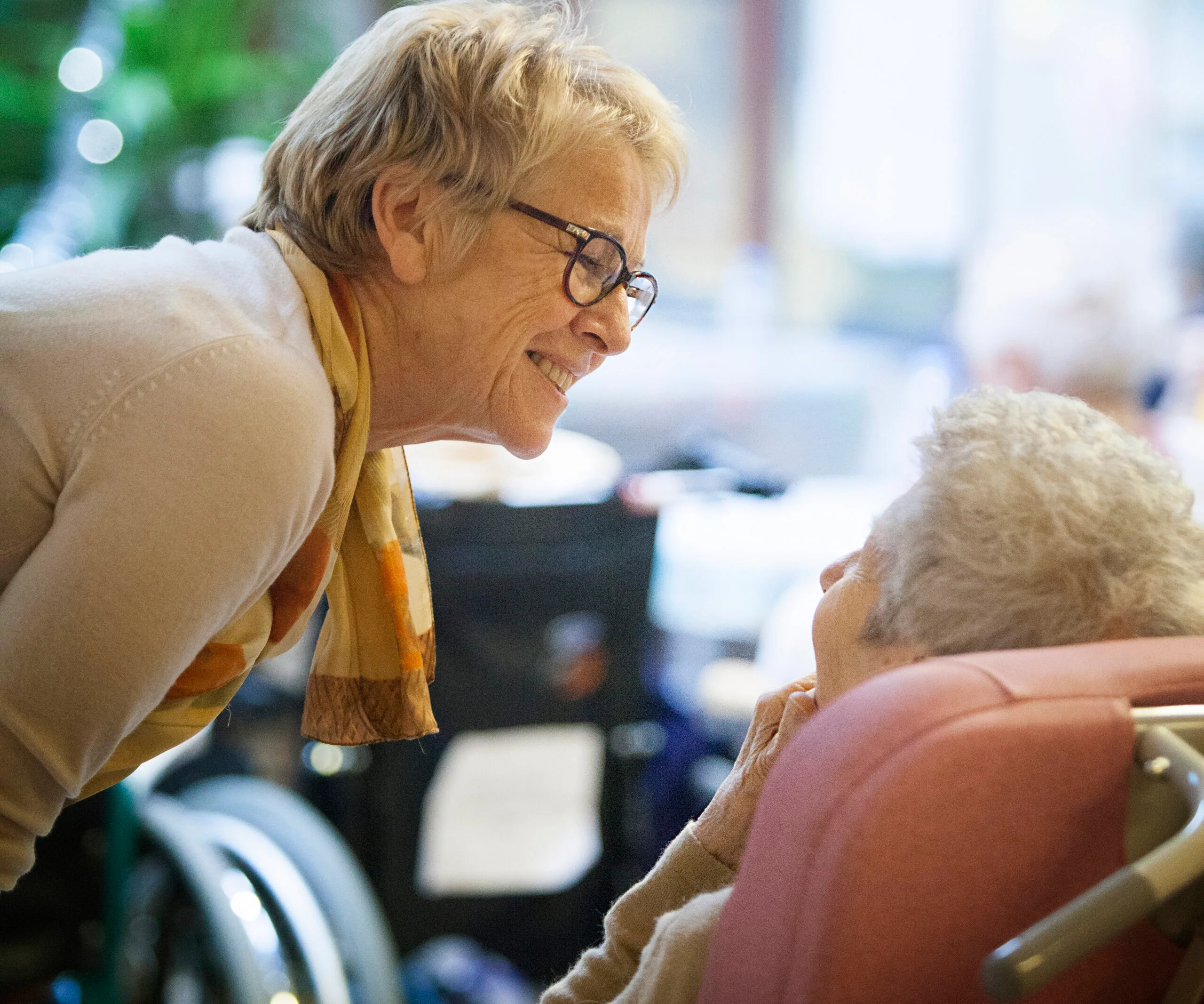 Elderly woman in glasses smiling at another older person in a care facility, showing warmth and connection.
