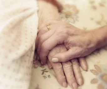 An elderly hand with a gold ring is gently held by another hand, symbolizing care and compassion.