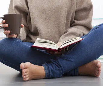 Person sitting cross-legged, holding a cup, reading a book with a red cover in casual attire.