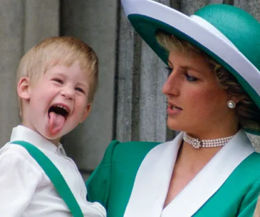 Young boy playfully sticks out tongue while being held by a woman in a green outfit and wide-brimmed hat.