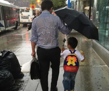 Father holds umbrella over son with lion backpack while walking in the rain.