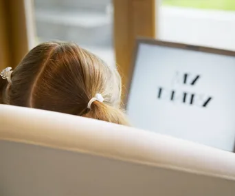 Girl with pigtails sitting on a chair, looking at a screen displaying blurred text.