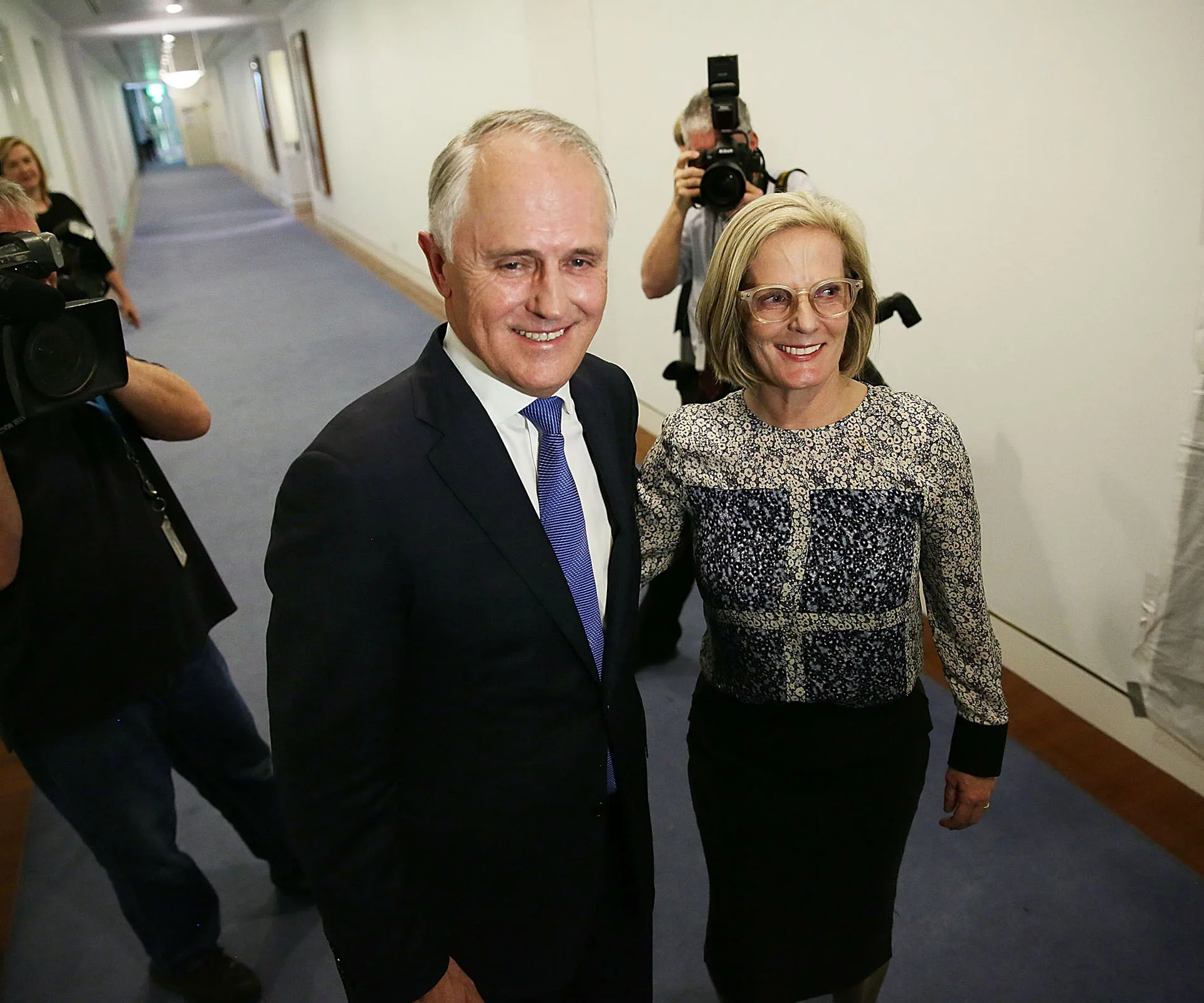 A man in a suit and a woman in glasses smile as they walk down a hallway, followed by photographers.